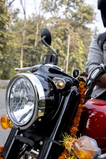 A close-up view of a black motorcycle, prominently featuring its front headlamp and handlebars. The motorcycle is adorned with marigold garlands and ribbons, suggesting a celebratory or festive context. A partial view of the rider's arm is visible, wearing a grey jacket. The background shows a blurred image of trees, indicating an outdoor setting.