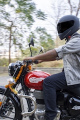 A person wearing a helmet rides a red Royal Enfield motorcycle, decorated with marigold garlands. The background is blurred, indicating motion, and shows green trees.