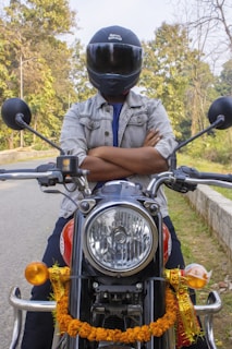 A person wearing a helmet is sitting on a motorcycle with arms crossed. The motorcycle is decorated with a garland and has a shiny headlight. The background includes a road lined with trees, indicative of a forest or park setting.