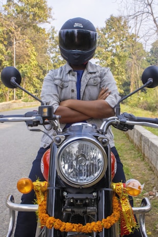 A person wearing a helmet is sitting on a motorcycle with arms crossed. The motorcycle is decorated with a garland and has a shiny headlight. The background includes a road lined with trees, indicative of a forest or park setting.