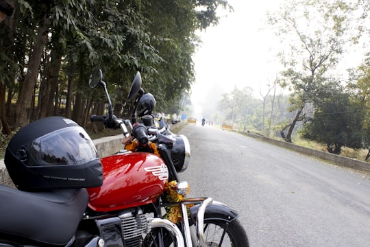 A parked motorcycle with a red fuel tank and black details is seen on the side of a road lined with trees. A helmet rests on the seat, and the road stretches into the distance with a few vehicles in the background, surrounded by greenery.