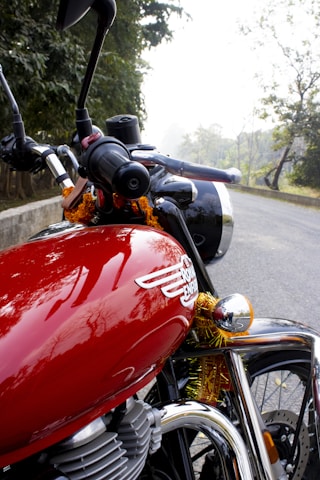 A close-up view of a shiny red motorcycle with a Royal Enfield badge, featuring chrome details and handlebars. The motorcycle is adorned with orange and yellow marigold garlands. It is parked on a road with lush green trees in the background, creating a serene outdoor setting.