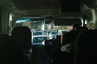 The image captures the inside of a vehicle, likely a bus, looking forward through the windshield. There are multiple people seated, visible from behind. The road ahead shows light traffic with several cars, and there is a visible stoplight indicating an intersection. The setting appears to be partially urban with hints of greenery.