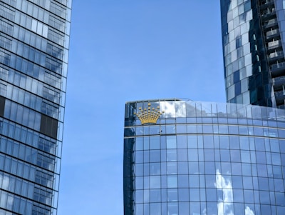 Tall, modern skyscrapers with reflective glass façades against a clear blue sky. A noticeable gold crown logo is seen on one of the buildings.
