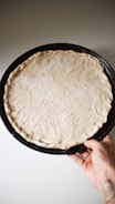 A hand holding a round baking tray with an uncooked pizza dough, lightly perforated and placed against a neutral background.