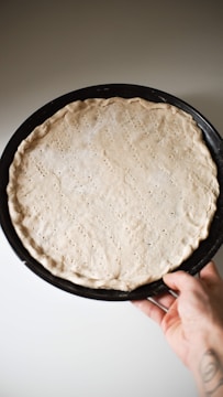A hand holding a round baking tray with an uncooked pizza dough, lightly perforated and placed against a neutral background.