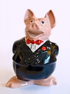 A ceramic figurine of a pig dressed in a formal black tuxedo with a red bow tie. The pig has a slightly exaggerated, cartoonish appearance with large eyes and a slight smile. The glossy surface reflects light, giving it a polished look.