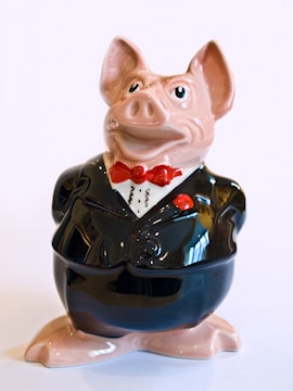 A ceramic figurine of a pig dressed in a formal black tuxedo with a red bow tie. The pig has a slightly exaggerated, cartoonish appearance with large eyes and a slight smile. The glossy surface reflects light, giving it a polished look.