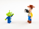 Two Lego minifigures are interacting with each other against a plain white background. One is a green alien with three eyes and a blue space suit, while the other is a human figure with a cowboy hat, a patterned shirt, and holding a small camera.