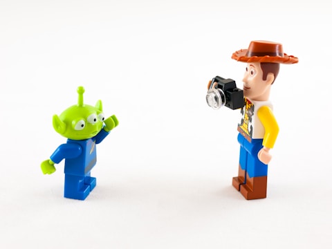Two Lego minifigures are interacting with each other against a plain white background. One is a green alien with three eyes and a blue space suit, while the other is a human figure with a cowboy hat, a patterned shirt, and holding a small camera.