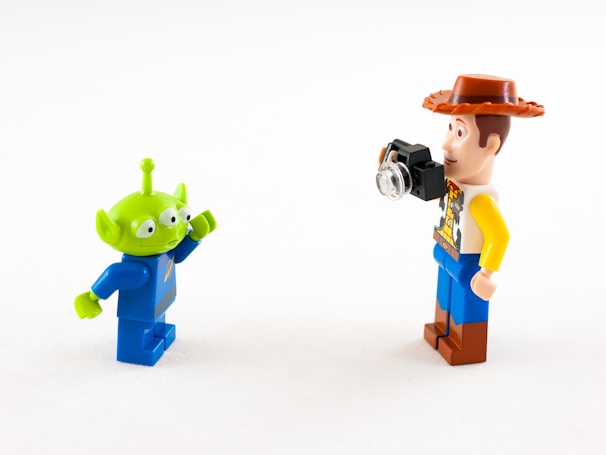Two Lego minifigures are interacting with each other against a plain white background. One is a green alien with three eyes and a blue space suit, while the other is a human figure with a cowboy hat, a patterned shirt, and holding a small camera.