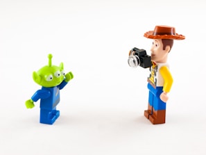 Two Lego minifigures are interacting with each other against a plain white background. One is a green alien with three eyes and a blue space suit, while the other is a human figure with a cowboy hat, a patterned shirt, and holding a small camera.
