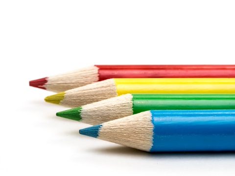 Four sharpened colored pencils are arranged in parallel, showcasing their distinct colors: red, yellow, green, and blue. The tips are pointed, highlighting the precision and readiness for use. The background is simple and white, emphasizing the vibrant hues of the pencils.