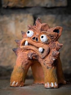 An earthy-colored sculpture with a rough texture, exhibiting exaggerated facial features with large, protruding white eyes, a flattened nose, and prominent teeth. The sculpture has four legs, each ending in a small, white-tipped foot. The background consists of a mottled, stone-like surface with varying shades of brown.