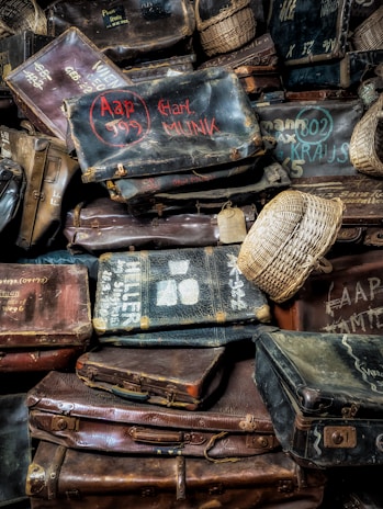 A pile of vintage leather suitcases with various sizes and shapes, showcasing signs of wear. Some have names and numbers painted on them in different colors, while a few woven baskets are interspersed among them.