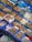 An assortment of seafood is displayed in a market setting. Various types of shellfish, octopus, and other sea creatures are organized in blue trays, with labels written in Korean. The seafood is fresh, with some items stored over ice or in shallow water.