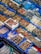 An assortment of seafood is displayed in a market setting. Various types of shellfish, octopus, and other sea creatures are organized in blue trays, with labels written in Korean. The seafood is fresh, with some items stored over ice or in shallow water.