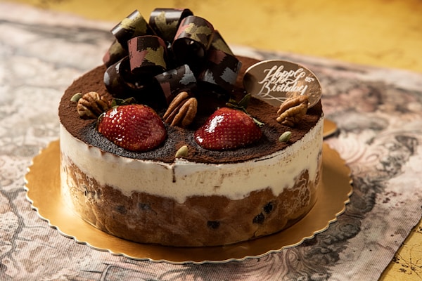 A beautifully decorated cake placed on a gold-colored platter. The cake has a creamy white frosting with chocolate shavings on top, garnished with fresh strawberries and pecan nuts. A large chocolate bow is centered on top, and a small chocolate plaque reads 'Happy Birthday.' The background is an elegant patterned tablecloth.