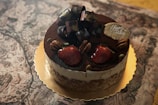 A beautifully decorated cake rests atop a golden base, featuring a top layer dusted with cocoa powder. Large, ripe strawberries and pecans are strategically placed to enhance its aesthetic appeal. A decorative bow and a small plaque with 'Happy Birthday' script add celebratory touches.