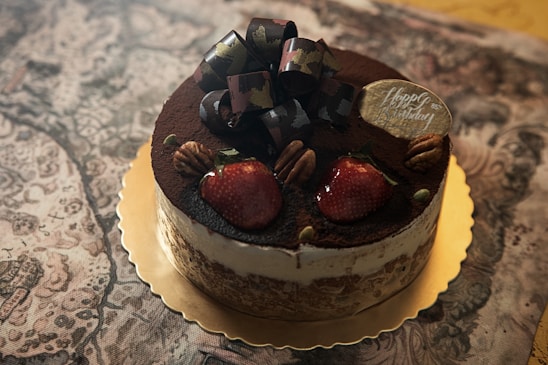 A beautifully decorated cake rests atop a golden base, featuring a top layer dusted with cocoa powder. Large, ripe strawberries and pecans are strategically placed to enhance its aesthetic appeal. A decorative bow and a small plaque with 'Happy Birthday' script add celebratory touches.
