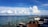 An outdoor seating area with wicker furniture and cushions overlooks a vast body of blue water under a bright sky filled with large white clouds. A few boats are seen on the horizon, and the water is dotted with buoys.