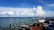 An outdoor seating area with wicker furniture and cushions overlooks a vast body of blue water under a bright sky filled with large white clouds. A few boats are seen on the horizon, and the water is dotted with buoys.