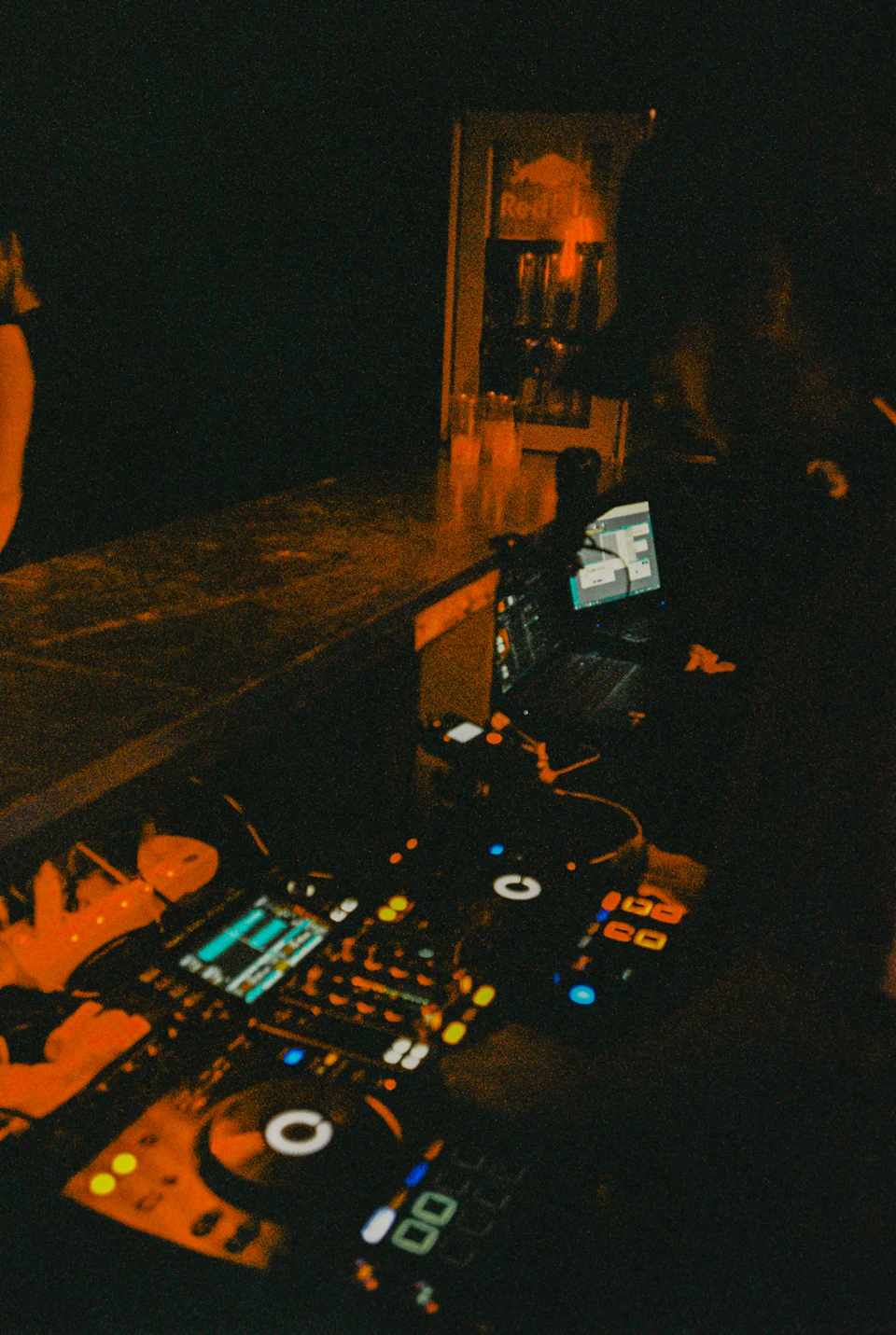 The image depicts a dimly lit room with a DJ booth at its center, featuring two turntables and a laptop displaying a video feed from another location. The turntables are illuminated by orange light, creating an atmospheric setting for music production or performance. In the background, there is a person partially visible on the right side of the image.