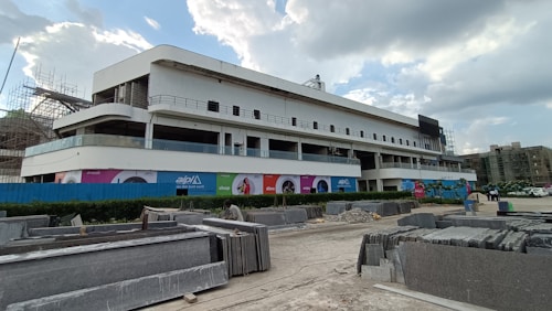 A modern, multi-story building under construction is centrally featured. The facade is primarily white, with large glass sections and several balconies. Surrounding the building are construction materials, including stacks of stone slabs. Advertising banners with colorful designs are visible on the building, and partly cloudy skies are overhead.