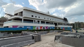 A modern, multi-story building under construction is centrally featured. The facade is primarily white, with large glass sections and several balconies. Surrounding the building are construction materials, including stacks of stone slabs. Advertising banners with colorful designs are visible on the building, and partly cloudy skies are overhead.