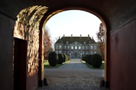 A grand estate is viewed through an arched gateway, with well-manicured shrubs lining the path leading to the house. The lighting casts intricate shadows on the walls of the archway, and the trees on either side display autumn colors.