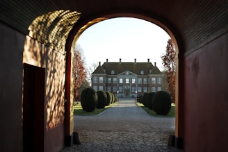 A grand estate is viewed through an arched gateway, with well-manicured shrubs lining the path leading to the house. The lighting casts intricate shadows on the walls of the archway, and the trees on either side display autumn colors.