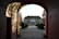 A grand estate is viewed through an arched gateway, with well-manicured shrubs lining the path leading to the house. The lighting casts intricate shadows on the walls of the archway, and the trees on either side display autumn colors.