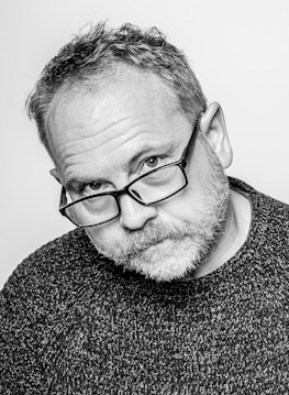 A black-and-white portrait of a middle-aged man with short hair and glasses. He has a thoughtful expression with a slight head tilt. The image is detailed, showing the texture of his sweater and facial hair.