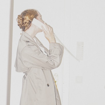 A person with braided hair is using a white landline telephone against a minimalist background. The person is wearing a beige coat and appears to be engaged in a conversation.