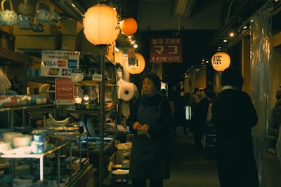 An indoor market scene features warm, ambient lighting from numerous hanging lanterns. Shelves filled with various items, including pottery and kitchenware, are visible. Several people walk and browse the aisles, creating a bustling yet cozy atmosphere.