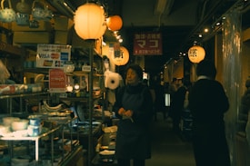 An indoor market scene features warm, ambient lighting from numerous hanging lanterns. Shelves filled with various items, including pottery and kitchenware, are visible. Several people walk and browse the aisles, creating a bustling yet cozy atmosphere.