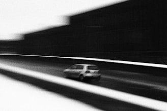 A blurred black and white image depicting a small car moving quickly on a road with a dynamic perspective. The surrounding environment and buildings appear indistinct due to the motion blur.