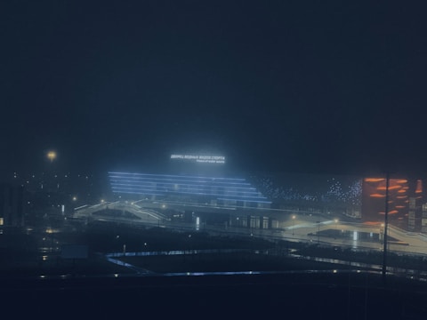 A modern building is illuminated against a dark night sky, featuring blue and orange LED lights. The sign on the building reads 'Palace of Water Sports' in both Cyrillic and English. The surrounding area is dimly lit, with streetlights providing additional illumination.