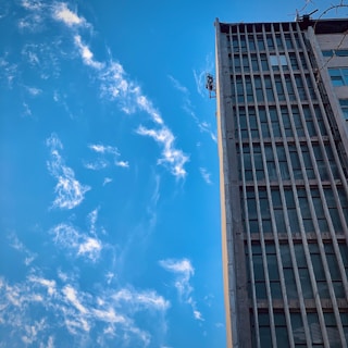 A modern building with a grid-like facade stands against a bright blue sky with scattered white clouds. A small platform or scaffold is attached to the side of the building, possibly for maintenance work.