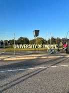 A large, prominent sign displaying the words 'University of Twente' beside a road. A bicycle is parked nearby on the sidewalk. In the background, there are trees and several flags waving in the distance under a clear blue sky.