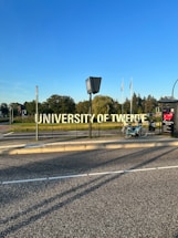 A large, prominent sign displaying the words 'University of Twente' beside a road. A bicycle is parked nearby on the sidewalk. In the background, there are trees and several flags waving in the distance under a clear blue sky.