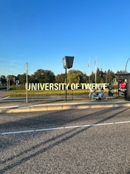 A large, prominent sign displaying the words 'University of Twente' beside a road. A bicycle is parked nearby on the sidewalk. In the background, there are trees and several flags waving in the distance under a clear blue sky.