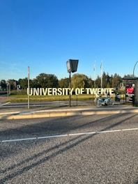 A large, prominent sign displaying the words 'University of Twente' beside a road. A bicycle is parked nearby on the sidewalk. In the background, there are trees and several flags waving in the distance under a clear blue sky.