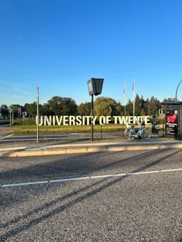 A large, prominent sign displaying the words 'University of Twente' beside a road. A bicycle is parked nearby on the sidewalk. In the background, there are trees and several flags waving in the distance under a clear blue sky.