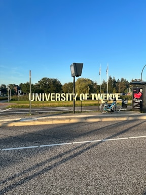 A large, prominent sign displaying the words 'University of Twente' beside a road. A bicycle is parked nearby on the sidewalk. In the background, there are trees and several flags waving in the distance under a clear blue sky.