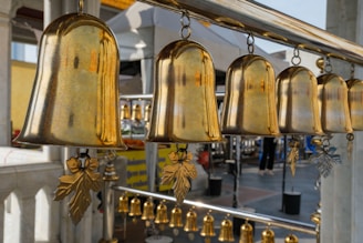 Several large, shiny brass bells hang in a row, each with ornate leaf-shaped pendants attached to their clappers. The background features an outdoor setting with marble columns and various smaller bells, contributing to a sense of tranquility and tradition.