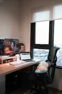A cozy office setup with a wooden desk featuring a computer monitor displaying a festive holiday scene, a laptop, a mechanical keyboard, and a calendar on the desk. A comfortable office chair with a cushion is positioned in front of the desk. A large window beside the desk lets in soft natural light.