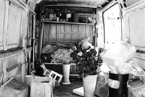 The interior of a van filled with various items, including potted plants, plastic containers, and rolls of paper or fabric. Shelves hold additional supplies and tools, while light spills in from an open door.