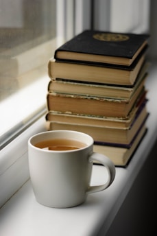 A white mug filled with tea is placed on a windowsill next to a stack of hardcover books. The books are piled unevenly, with some wear on their pages and covers. The scene is lit by natural light coming through the window.
