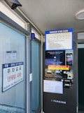A parking payment kiosk with a screen displaying information in Korean. The kiosk includes a payment terminal and various instructions. Adjacent to the kiosk is a glass window with signage about parking fees and discounts. An overhead security camera is visible.